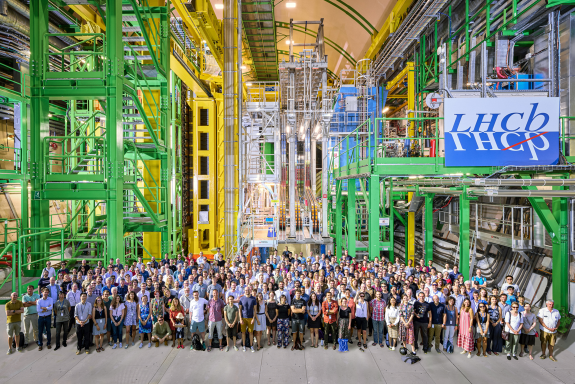 The LHCb collaboration in front of the LHCb Upgrade 1 detector in the underground cavern. The structures in green, yellow, grey and blue are used to support the different detectors